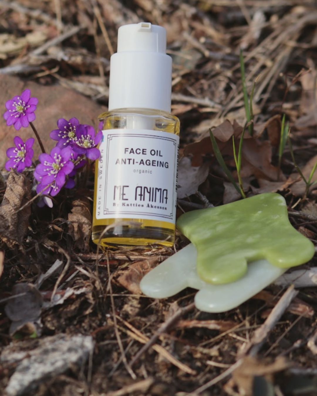 Me Anima Face Oil Anti-Ageing placerad på marken bland löv och blommor bredvid två gröna Gua Sha-verktyg